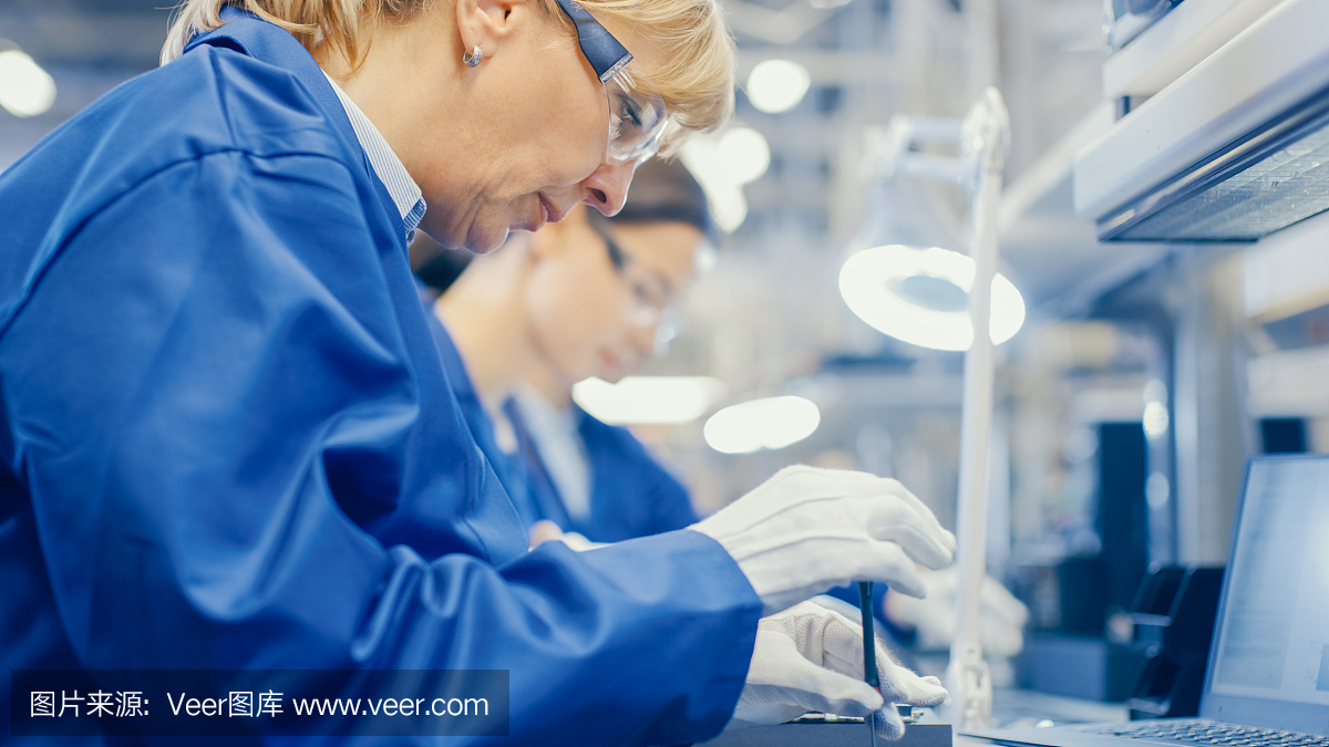 电子工厂的女工穿着蓝色工作服,戴着防护眼镜,正在用螺丝刀组装智能手机。高科技工厂设施与更多的员工在后台。